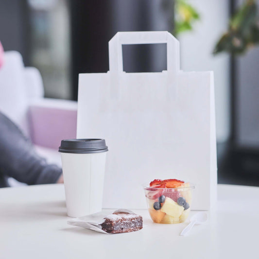 White paper bag with flat handles next to coffee cup, fruit salad, and brownie on a table.